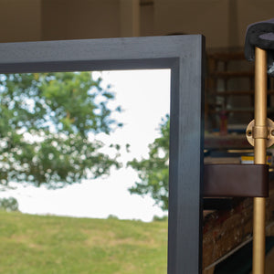 The corner of a mirror in a grey-blue timber frame. The glass is reflecting the East Sussex countryside. On the right-hand side, a brown leather strap attaches an antique brass pole to the mirror frame. 