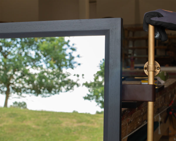 The corner of a mirror in a grey-blue timber frame. The glass is reflecting the East Sussex countryside. On the right-hand side, a brown leather strap attaches an antique brass pole to the mirror frame. 
