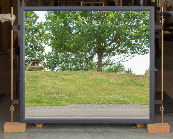 A large rectangular mirror with a slate-blue timber-stained frame and leather straps supporting two antique brass poles leaning against in a bench in our workshop. The mirror is reflecting the East Sussex countryside. 