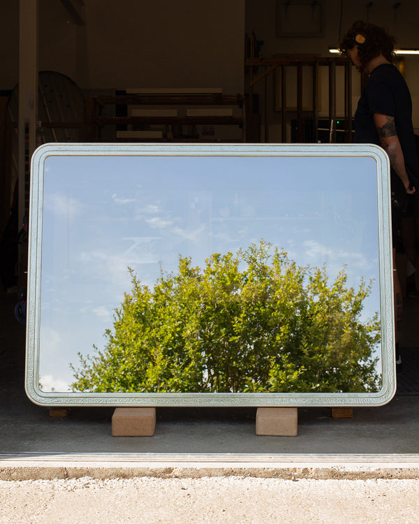 A bespoke rectangular TV mirror with rounded corners leaning against a bench in a workshop with a person working at the bench behind. The frame is finished in gilded white gold with a beaten white gold section in the centre. The dielectric mirror glass is reflecting the top of a tree and a blue sky. 