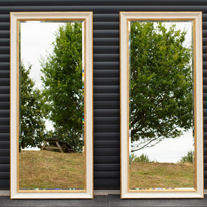 two tall mirrors standing side-by-side against a grey door. The frames are finished in an aged cracked gesso with antique gold detailing and the mirror has a bevel along the outside and is reflecting the East Sussex countryside.
