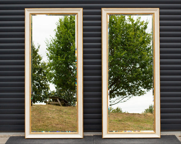two tall mirrors standing side-by-side against a grey door. The frames are finished in an aged cracked gesso with antique gold detailing and the mirror has a bevel along the outside and is reflecting the East Sussex countryside.
