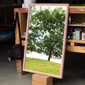 A rectangular mirror painted light pink with a round, bobbin detail that runs along the entire length of the face of the frame. The mirror is leaning against a bench in our workshop and the mirror glass is reflecting the East Sussex countryside