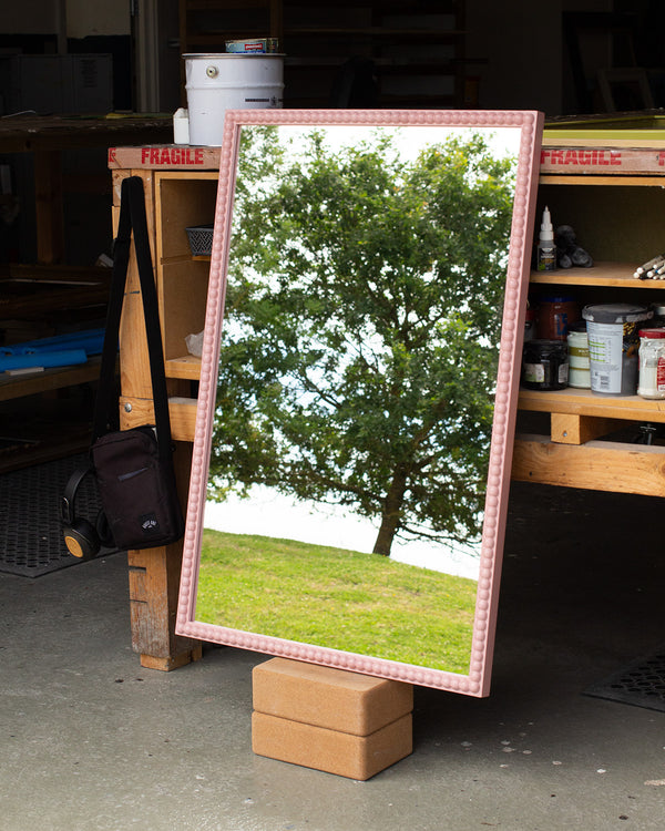 A rectangular mirror painted light pink with a round, bobbin detail that runs along the entire length of the face of the frame. The mirror is leaning against a bench in our workshop and the mirror glass is reflecting the East Sussex countryside