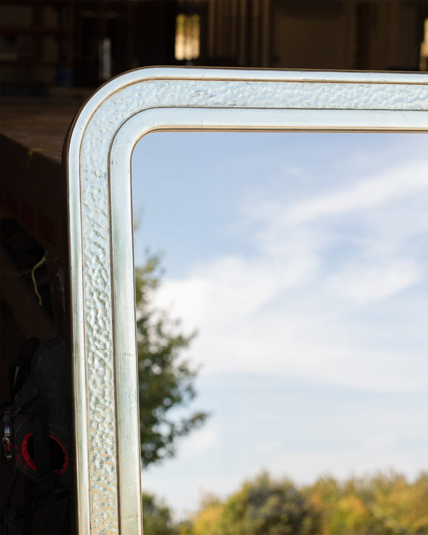A close-up of a rounded corner of a bespoke TV mirror finished in gilded white gold with a beaten section in the centre. The dielectric mirror glass is reflecting the sky and some trees. 