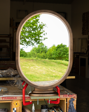 A shaped mirror with a bronze frame secured to a bench in our workshop with red clamps. The mirror is supported by a tiered base so it can be secured to a  flat surface. 