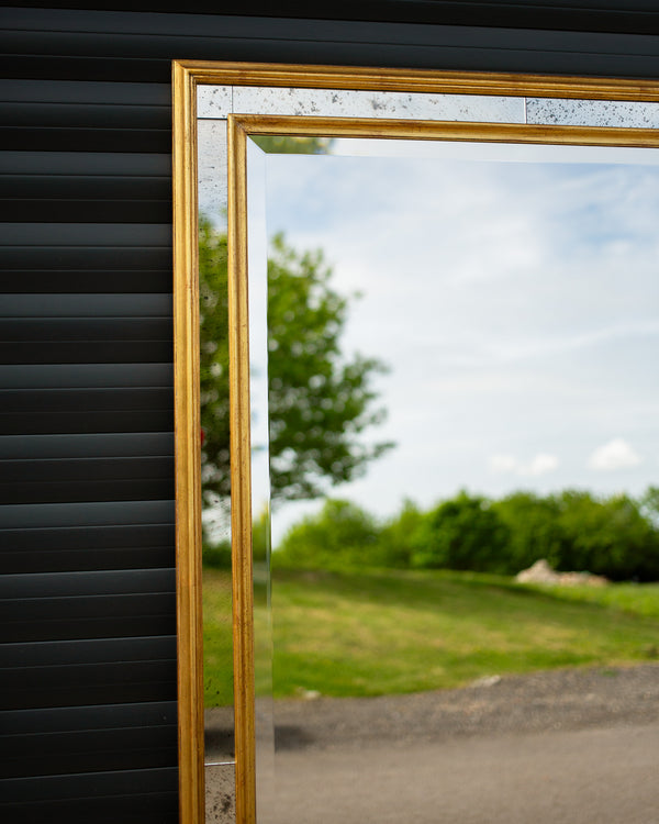The corner detail of a bespoke mirror made using an antique gold frame with a Venetian mirror inlay. The central mirror has a bevel and is reflecting the East Sussex countryside. This traditional style mirror is leaning against a workshop roller door. 