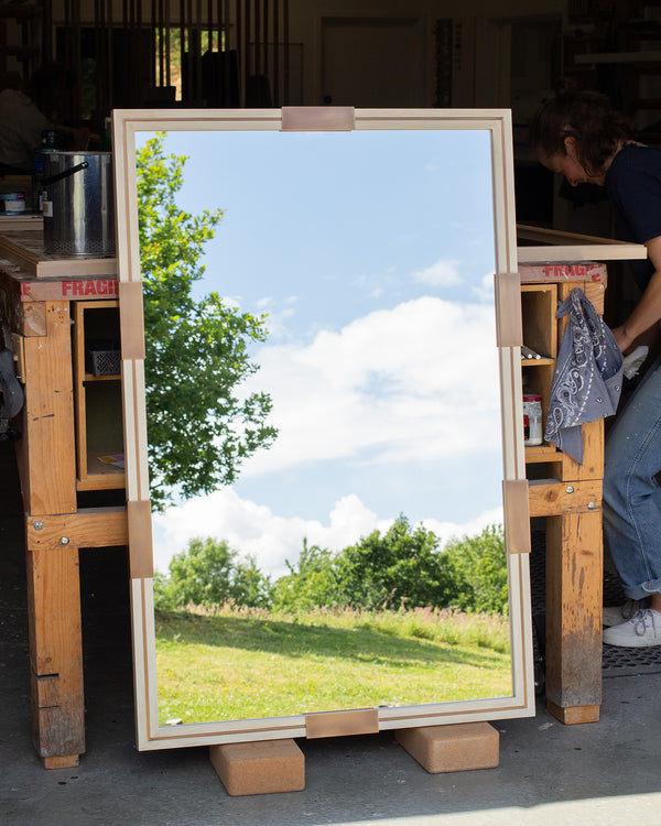 A distressed white mirror with an antique brass inlay running through the centre of the frame and antique brass clips along the edges. The mirror is propped against a bench in a workshop and the mirror glass is reflecting the East Sussex country. 