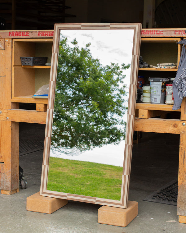 A narrow rectangular mirror leaning against a workbench. The frame is finished in a walnut and light grey sycamore chequered design, and the mirror glass is reflecting a grassy hill and a tree outside our East Sussex workshop. 