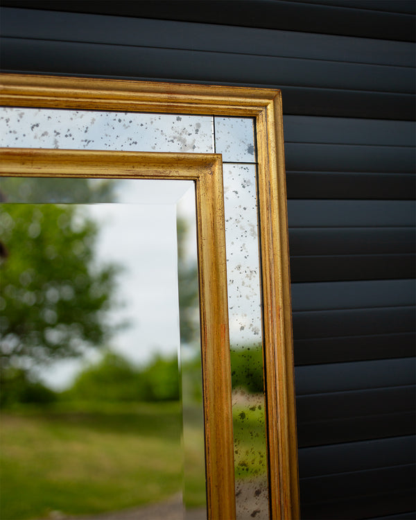 The corner detail of a bespoke mirror made using an antique gold frame with a Venetian mirror inlay. The central mirror has a bevel and is reflecting the East Sussex countryside. This traditional style mirror is leaning against a workshop roller door. 
