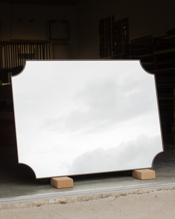 A large bespoke mirror with scallop corners leaning against a workbench in our workshop. The frame of the mirror is made using a slim 6mm wide bronze metal tray frame, and the mirror glass is reflecting an overcast grey sky. 