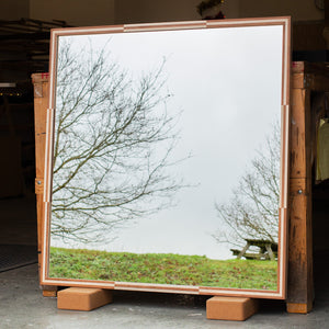 A square mirror leaning against a workbench. The mirror frame is finished in a cherry and pink burr veneer pattern. The mirror glass is reflecting the East Sussex countryside and a picnic bench. 