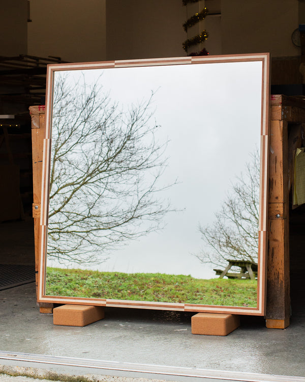 A square mirror leaning against a workbench. The mirror frame is finished in a cherry and pink burr veneer pattern. The mirror glass is reflecting the East Sussex countryside and a picnic bench. 