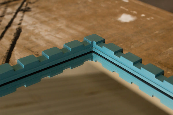 The corner detail of a mirror that is laying flat on a paper-covered work bench. The frame profile is made using a Dentil moulding with raised rectangles that run along the length. The frame is hand-painted using Little Greene's Tea. 