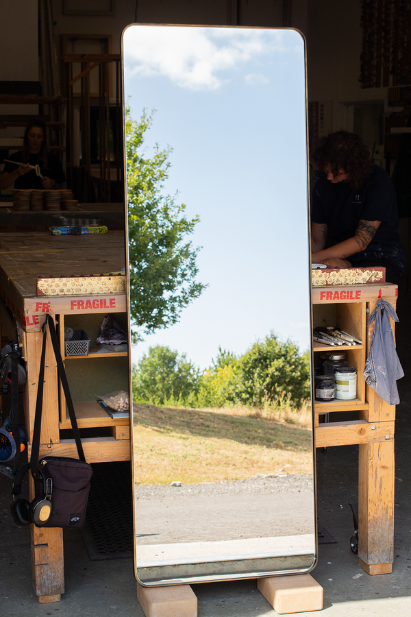 A tall and narrow mirror made with a thin 6mm wide tray frame finished in our imitation antique brass. The is leaning against a bench in our workshop and the mirror glass is reflecting the East Sussex countryside outside our workshop. 