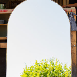An archtop mirror with a thin imitation brass frame measuring 6mm wide. The mirror glass is reflecting a blue sky and the top of a tree. 