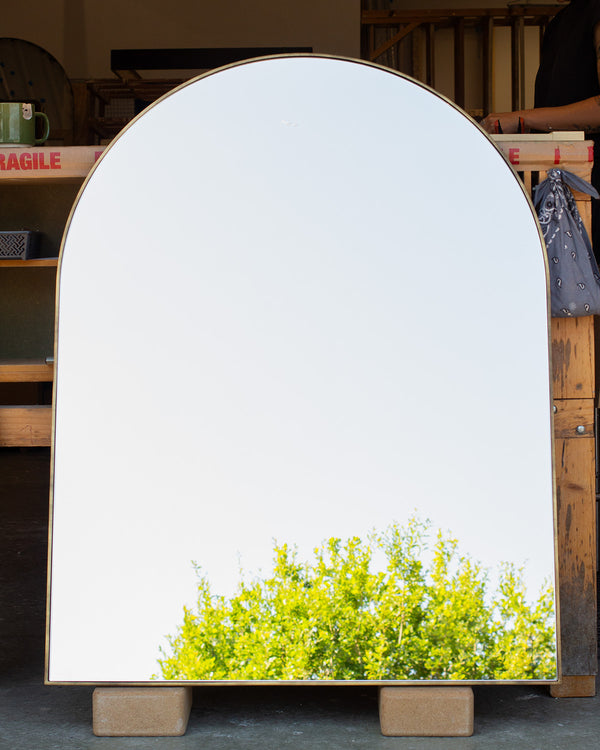An archtop mirror with a thin imitation brass frame measuring 6mm wide. The mirror glass is reflecting a blue sky and the top of a tree. 