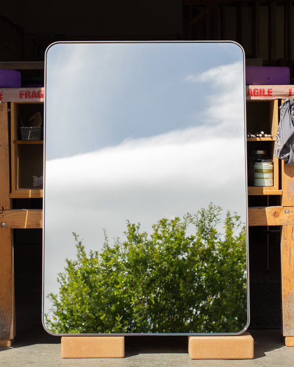 A rectangular mirror with radial coerns leaning against a bench in our wood workshop. The mirror's frame is a thin 6mm wide and is made of polished stainless steel. The mirror glass is reflecting a cloudy sky and the top of a tree. 
