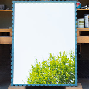 A bespoke teal-coloured mirror. The frame is made using a dentil profile, which has raised rectangular sections evenly spaced along the frame, which is painted Little Greene's Tea with a satin finish. The mirror glass is reflecting the sky and the top of a tree. 