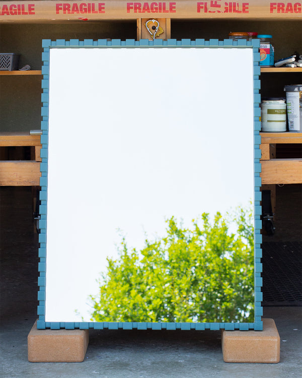 A bespoke teal-coloured mirror. The frame is made using a dentil profile, which has raised rectangular sections evenly spaced along the frame, which is painted Little Greene's Tea with a satin finish. The mirror glass is reflecting the sky and the top of a tree. 