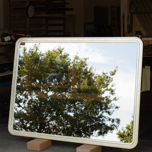 A large bespoke TV mirror leaning against a bench in a workshop. The frame is finished in gilded white gold with a beaten white gold section in the centre. The mirror is made of dielectric mirror glass and is partially transparent and partially reflecting a tree and bit of light blue sky. 