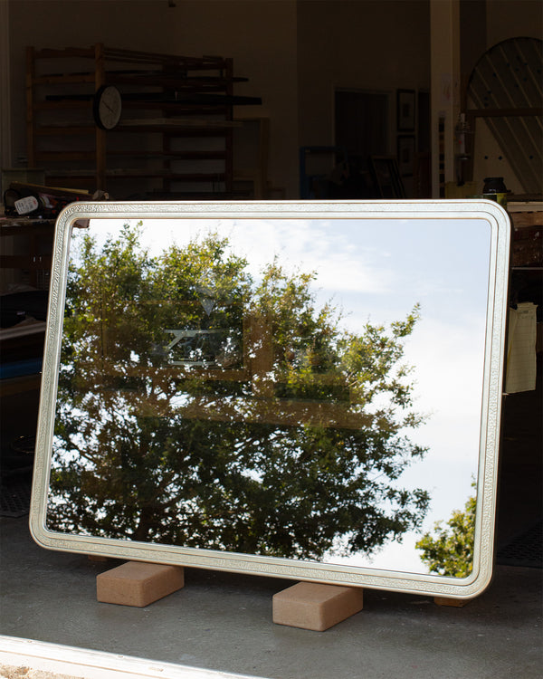 A large bespoke TV mirror leaning against a bench in a workshop. The frame is finished in gilded white gold with a beaten white gold section in the centre. The mirror is made of dielectric mirror glass and is partially transparent and partially reflecting a tree and bit of light blue sky. 