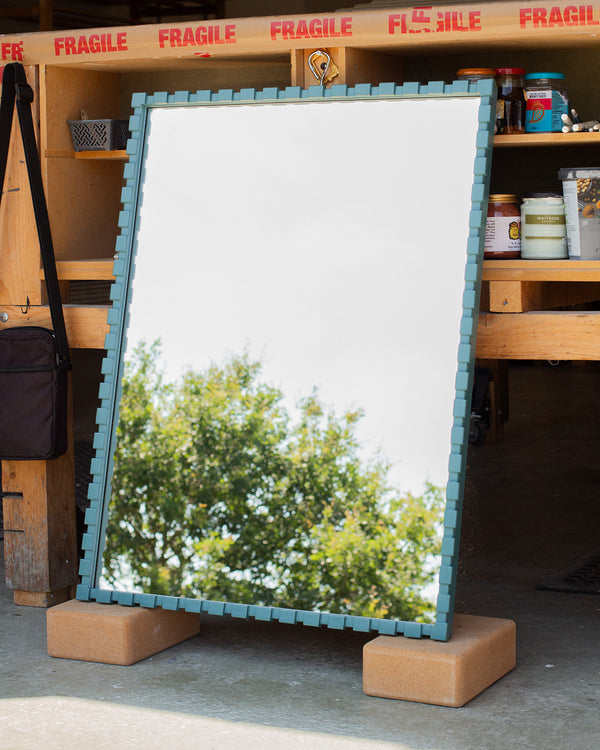 The side view of a hand-painted bespoke teal-coloured mirror. The frame is made using a dentil profile, which has raised rectangular sections evenly spaced along the frame, which is painted Little Greene's Tea with a satin finish. The mirror glass is reflecting the sky and the top of a tree. 