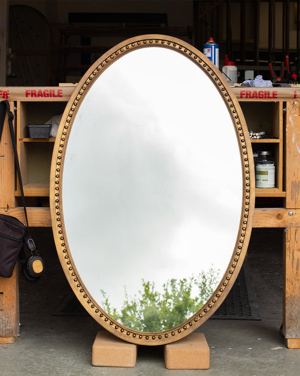 An oval mirror leaning against a work bench in a workshop. The frame is finished in an imitation brass and a bobbin detail runs along the inside of the frame. The mirror glass is lightly antiqued with heavier antiquing along the edges. The mirror is reflecting a grey sky and part of a tree. 