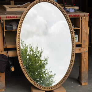 A photo taken from the side of a oval mirror in our workshop. The frame is finished in an imitation brass and a bobbin detail runs along the inside of the frame. The mirror glass is lightly antiqued with heavier antiquing along the edges and the mirror glass is reflecting the East Sussex countryside. 