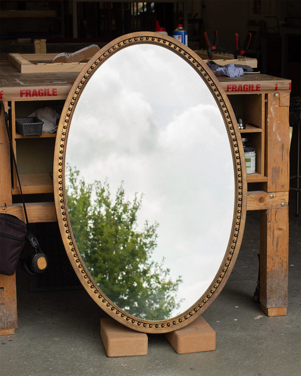 A photo taken from the side of a oval mirror in our workshop. The frame is finished in an imitation brass and a bobbin detail runs along the inside of the frame. The mirror glass is lightly antiqued with heavier antiquing along the edges and the mirror glass is reflecting the East Sussex countryside. 