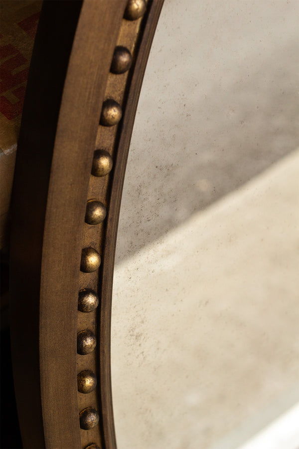 A close-up photo of an oval shaped mirror. The frame is finished in an imitation brass and a bobbin detail runs along the inside of the frame. The mirror glass is lightly antiqued with heavier antiquing along the edges. 