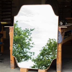 A large bespoke metal shaped mirror reflecting the East Sussex countryside. The metal finish is antique brass and the frame is thin and elegant. 