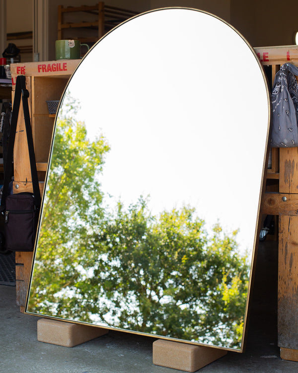 An archtop mirror with a thin 6mm wide frame finished in imitation brass leaning against a bench in our workshop. The mirror glass is reflecting a clear sky and the top of some trees. 