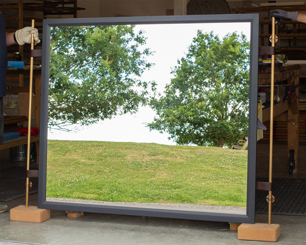 A side-view of a large rectangular mirror with a slate-blue timber-stained frame and leather straps supporting two antique brass poles leaning against in a bench in our workshop. The mirror is reflecting the East Sussex countryside. 