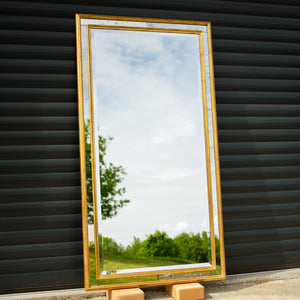 A bespoke mirror made using an antique gold frame with a Venetian mirror inlay. The central mirror has a bevel and is reflecting the East Sussex countryside. This traditional style mirror is leaning against a workshop roller door. 