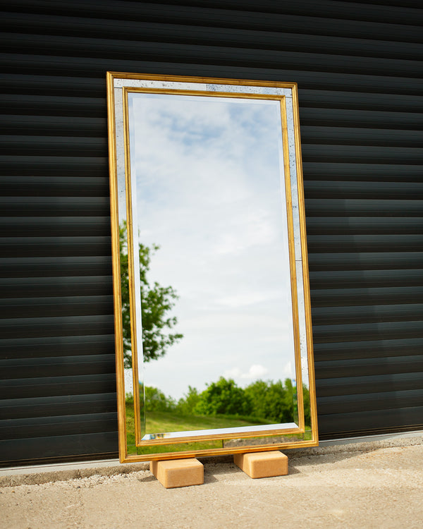 A bespoke mirror made using an antique gold frame with a Venetian mirror inlay. The central mirror has a bevel and is reflecting the East Sussex countryside. This traditional style mirror is leaning against a workshop roller door. 