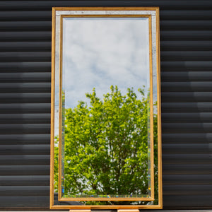 A bespoke mirror made using an antique gold frame with a Venetian mirror inlay. The central mirror has a bevel and is reflecting the East Sussex countryside. This traditional style mirror is leaning against a workshop roller door. 