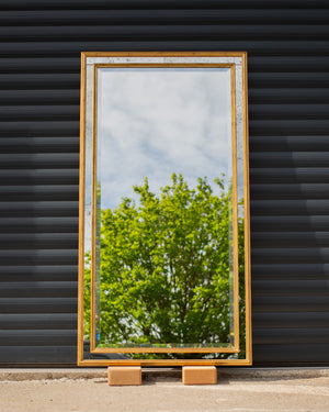 A bespoke mirror made using an antique gold frame with a Venetian mirror inlay. The central mirror has a bevel and is reflecting the East Sussex countryside. This traditional style mirror is leaning against a workshop roller door. 
