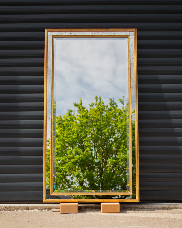 A bespoke mirror made using an antique gold frame with a Venetian mirror inlay. The central mirror has a bevel and is reflecting the East Sussex countryside. This traditional style mirror is leaning against a workshop roller door. 