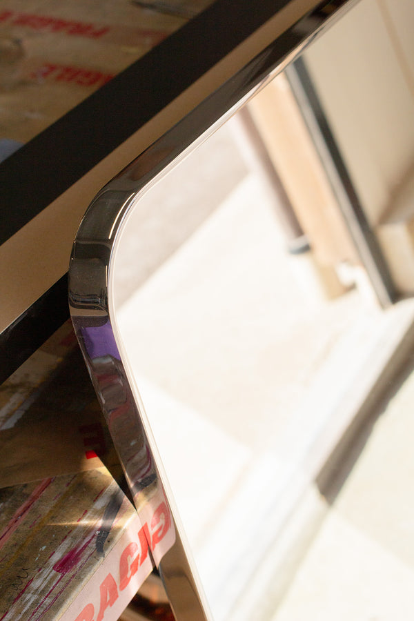 A close-up view of a rounded mirror corner. The slim frame of the mirror is made of polished stainless steel, 